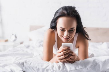 Akıllı telefon kullanarak yatakta gülümseyen esmer bir kadın.