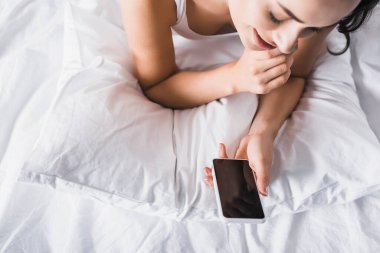 Yatakta elinde akıllı telefon olan esmer bir kadın.