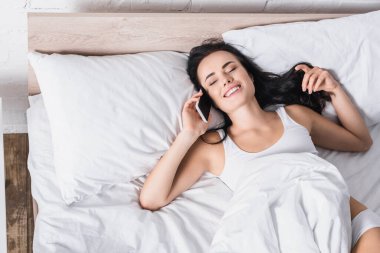 Gülümseyen genç esmer kadın yatakta akıllı telefonla konuşuyor.