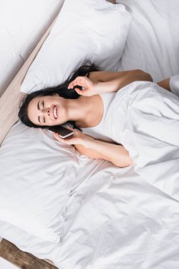 top view of happy young brunette woman talking on smartphone in bed clipart