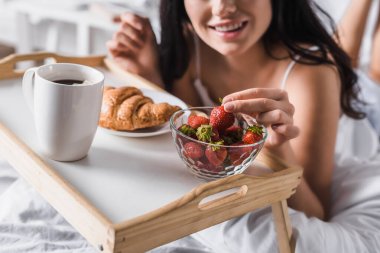 cropped view of young brunette woman having croissant, strawberry and cocoa for breakfast in bed clipart