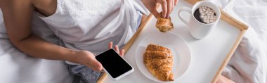 Kahvaltıda kruvasan ve kakao içen bir kadının yatakta akıllı telefon ve afiş kullanması görüntüsü.