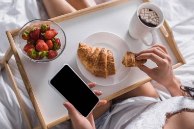 Kahvaltıda kruvasan, çilek ve kakao yiyen bir kadının yatakta akıllı telefon tutarken görüntüsü.