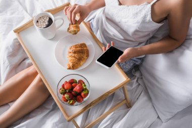 Kahvaltıda kruvasan, çilek ve kakao yiyen bir kadının yatakta akıllı telefon tutarken görüntüsü.