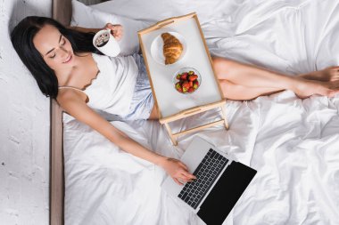 Genç esmer kadın laptopunu kullanırken yatakta kahvaltı yapıyor.