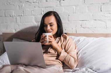 young brunette woman sitting in bed with mug and laptop at morning clipart
