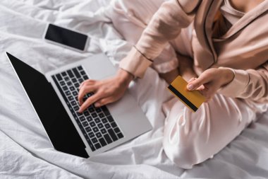 Akıllı telefon, kredi kartı ve dizüstü bilgisayarla yatakta oturan bir kadın görüntüsü.