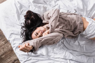 top view of smiling young brunette woman in sweater lying in bed at morning clipart