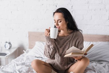 young brunette woman in sweater sitting in bed with mug of hot cocoa and book at morning clipart