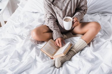 cropped view of woman in sweater and knitted socks sitting in bed with mug of hot cocoa and book at morning clipart