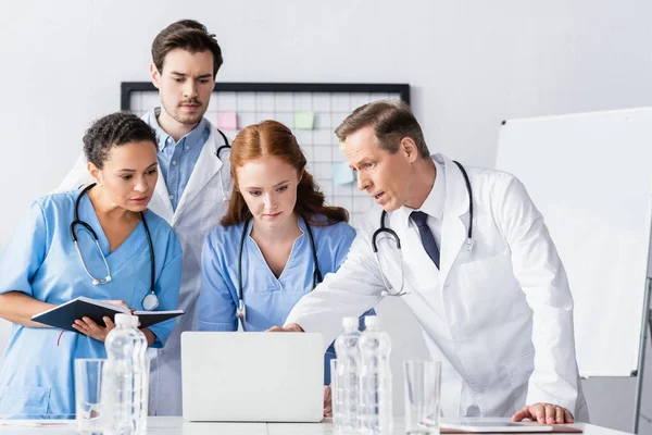 Multiethnic doctors and nurses with notebook looking at laptop near water on blurred foreground