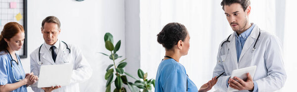 African american nurse standing near doctor with digital tablet and colleagues with laptop in clinic, banner 