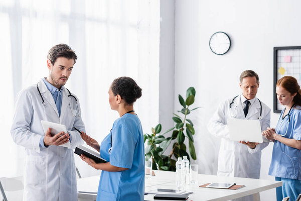 Многонациональный персонал больницы с цифровым планшетом и ноутбуком разговаривает рядом с коллегами с ноутбуком на размытом фоне в клинике 