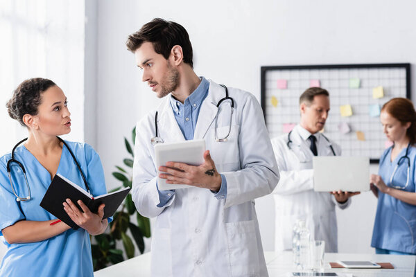 Multiethnic hospital staff with notebook and digital tablet talking near colleagues with laptop on blurred background 