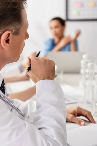 Médico Con Pluma Cuaderno Sentado Cerca Colegas Que Trabajan Fondo — Foto de Stock