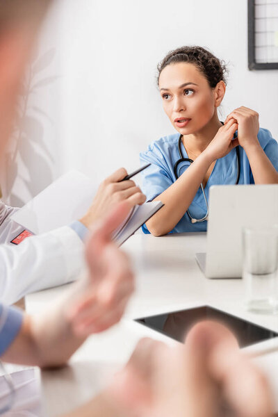 Африканская американская медсестра смотрит на коллегу с ноутбуком рядом с устройствами на размытом переднем плане на столе 