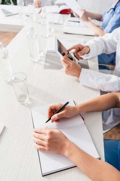 Обрезанный вид медсестры, пишущей на ноутбуке рядом с врачом с цифровыми таблетками и бутылками воды на размытом фоне 