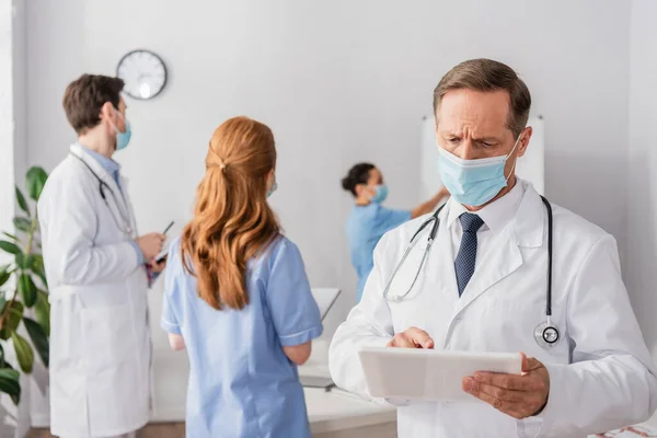 Tıbbi Maskeli Doktor Arka Planda Bulanık Etnik Meslektaşlarıyla Dijital Tablet — Stok fotoğraf