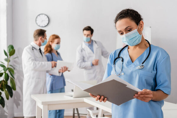 African american nurse in medical mask looking at paper folder with blurred colleagues working on background