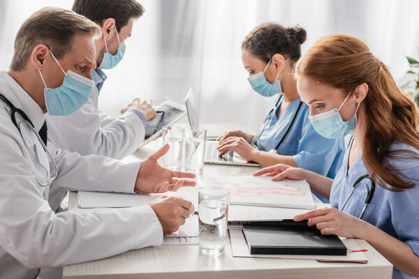 Multiethnic hospital staff working while sitting at workplace with papers, devices and glasses of water in hospital