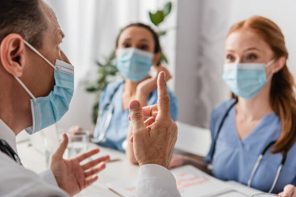 Doctor in medical mask pointing with finger while sitting at workplace with blurred multiethnic colleagues on background