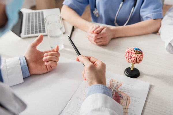 Cropped view of doctor with pen gesturing near colleagues at workplace with papers and brain anatomical model on blurred foreground
