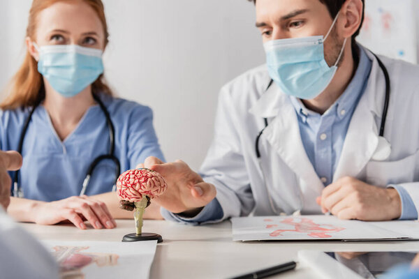 Doctor in medical mask touching brain anatomical model while sitting at workplace near colleagues on blurred foreground