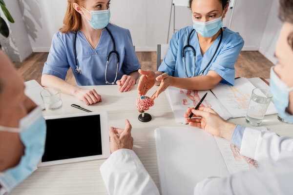 African american nurse pointing with hands at brain anatomical model near colleagues at workplace on blurred foreground