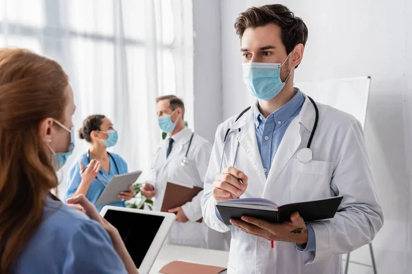 Doctor in medical mask holding notebook while talking to nurse with blurred multicultural colleagues on background