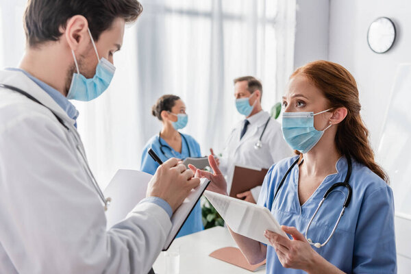 Redhead nurse talking to doctor writing in notebook with blurred multicultural colleagues on background