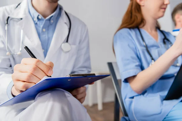 在模糊的背景下 坐在护士身边时 医生在剪贴板上用听诊器写字的剪影 — 图库照片