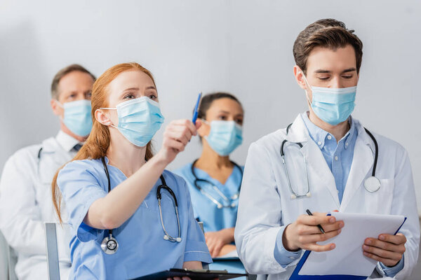 Redhead nurse pointing with pen while sitting near doctor during meeting with blurred colleagues on background in hospital