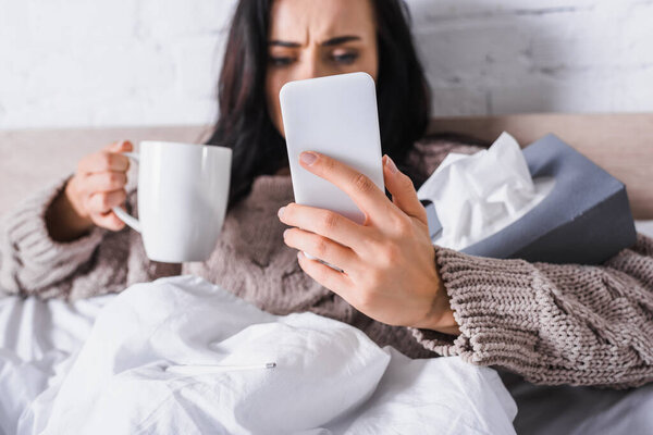 sick young brunette woman with hot drink and tissue holding smartphone in bed, blurred background