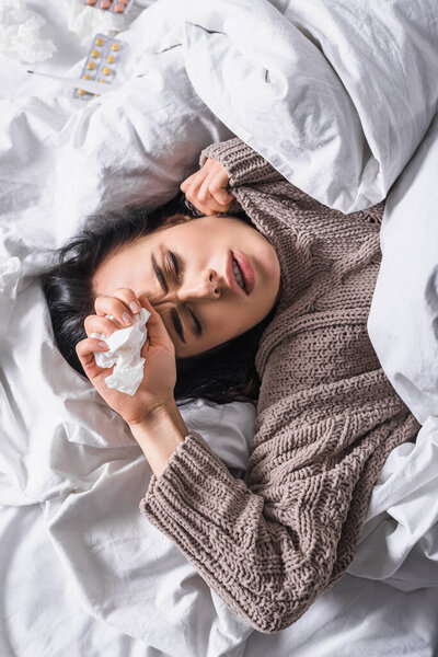 top view of sick young brunette woman with pills and tissue in bed