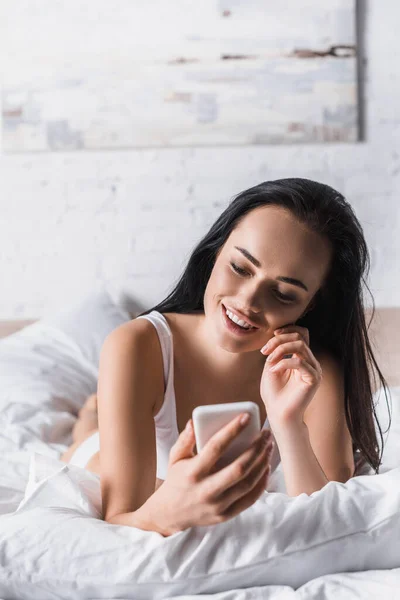 Χαμογελαστή Νεαρή Μελαχρινή Γυναίκα Στο Κρεβάτι Χρησιμοποιώντας Smartphone — Φωτογραφία Αρχείου