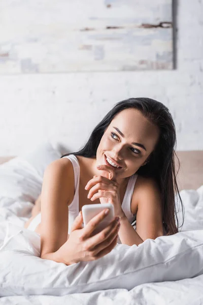夢のような笑顔若いブルネット女性でベッドでスマートフォンを使用して — ストック写真
