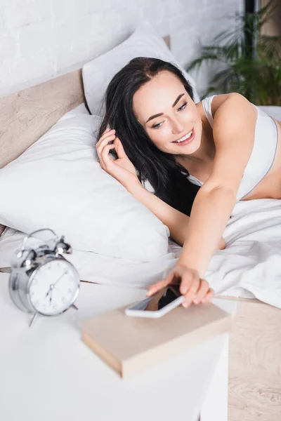 若いブルネットの女性が目覚まし時計の近くにスマートフォンを置くベッドで — ストック写真