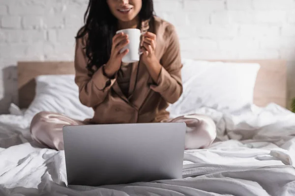 若いブルネットの女性が朝にマグカップとラップトップでベッドに座っているのをクロップドビュー — ストック写真