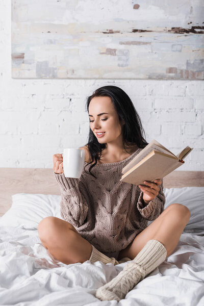 smiling young brunette woman in sweater sitting in bed with mug of hot cocoa and book at morning