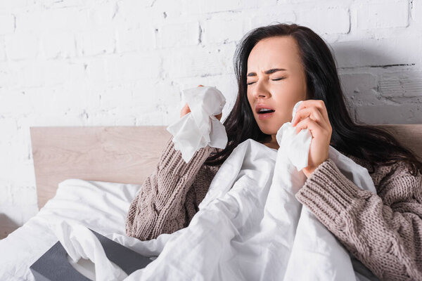 ill young brunette woman in sweater sneezing in bed with tissue