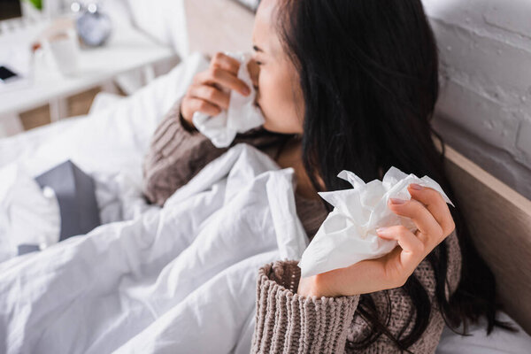 blurred ill young brunette woman in sweater sneezing in bed with tissue