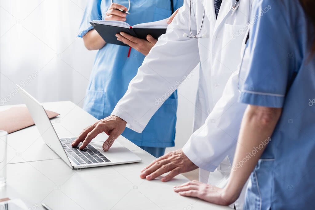 Cropped view of doctor in white coat using laptop near nurses with notebook 