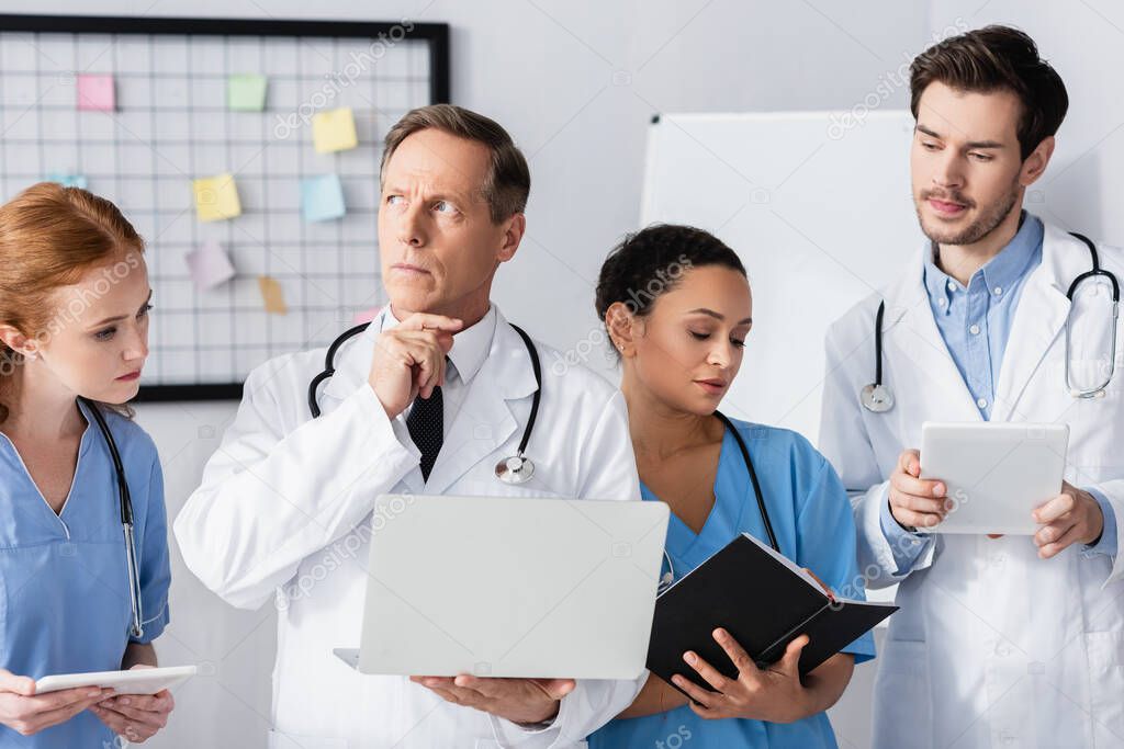 Pensive doctor with laptop standing near multiethnic colleagues in hospital 