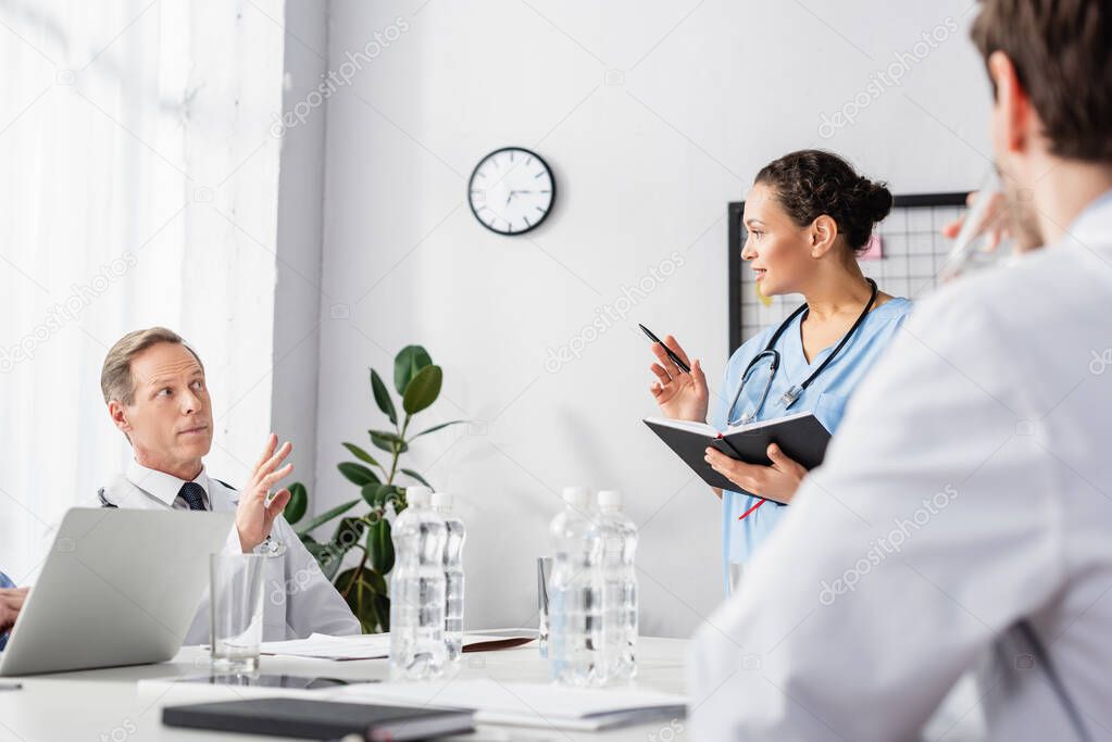 African american nurse with notebook and pen looking at doctor with raised hand near colleague on blurred foreground 