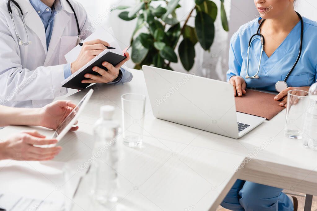 Cropped view of multiethnic hospital staff using devises while working with notebook and paper folder near bottles of water on blurred foreground 