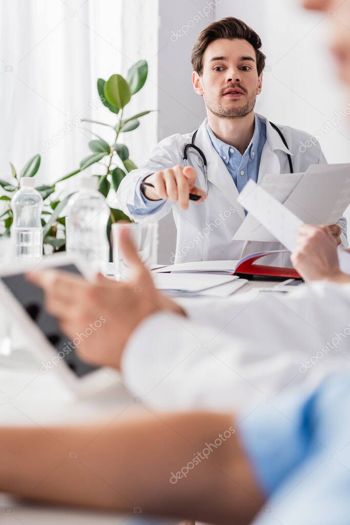 Doctor with papers pointing with finger near colleagues with digital tablet on blurred foreground 