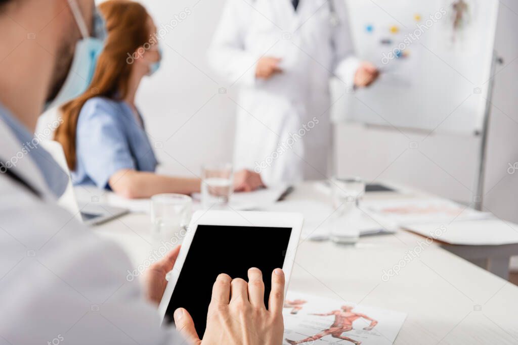 Doctor in medical mask using digital tablet with blank screen near document and colleagues on blurred background 