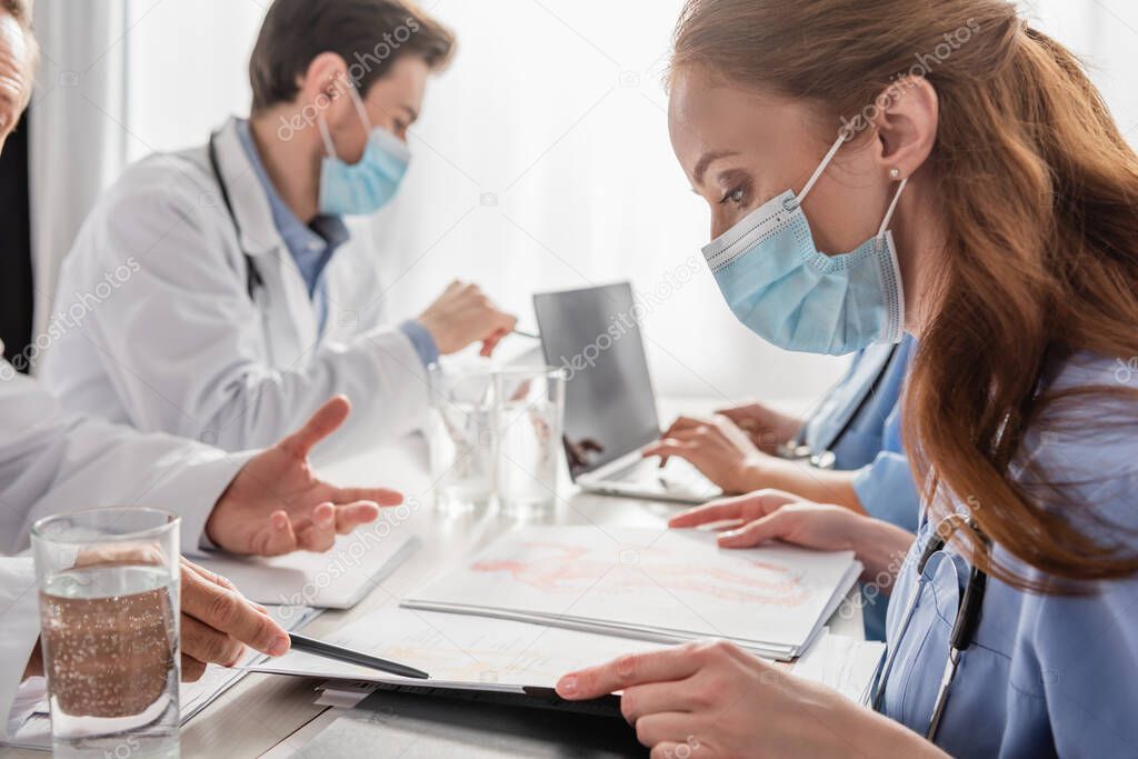 Redhead nurse looking at paper folder with pictures while sitting near doctor pointing with pen near colleagues on blurred background