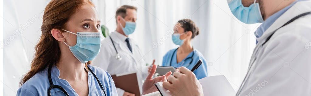 Redhead nurse talking to doctor writing in notebook with blurred multicultural colleagues on background, banner