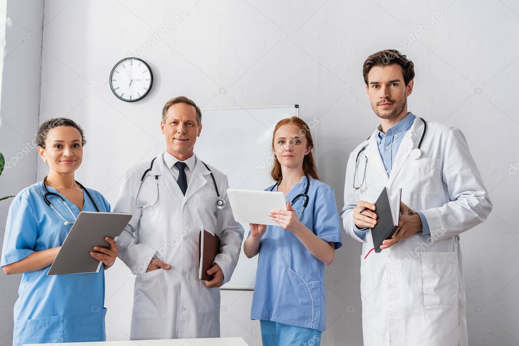 Positive multiethnic hospital staff with papers and digital tablet looking at camera with flipchart and wall clock on background